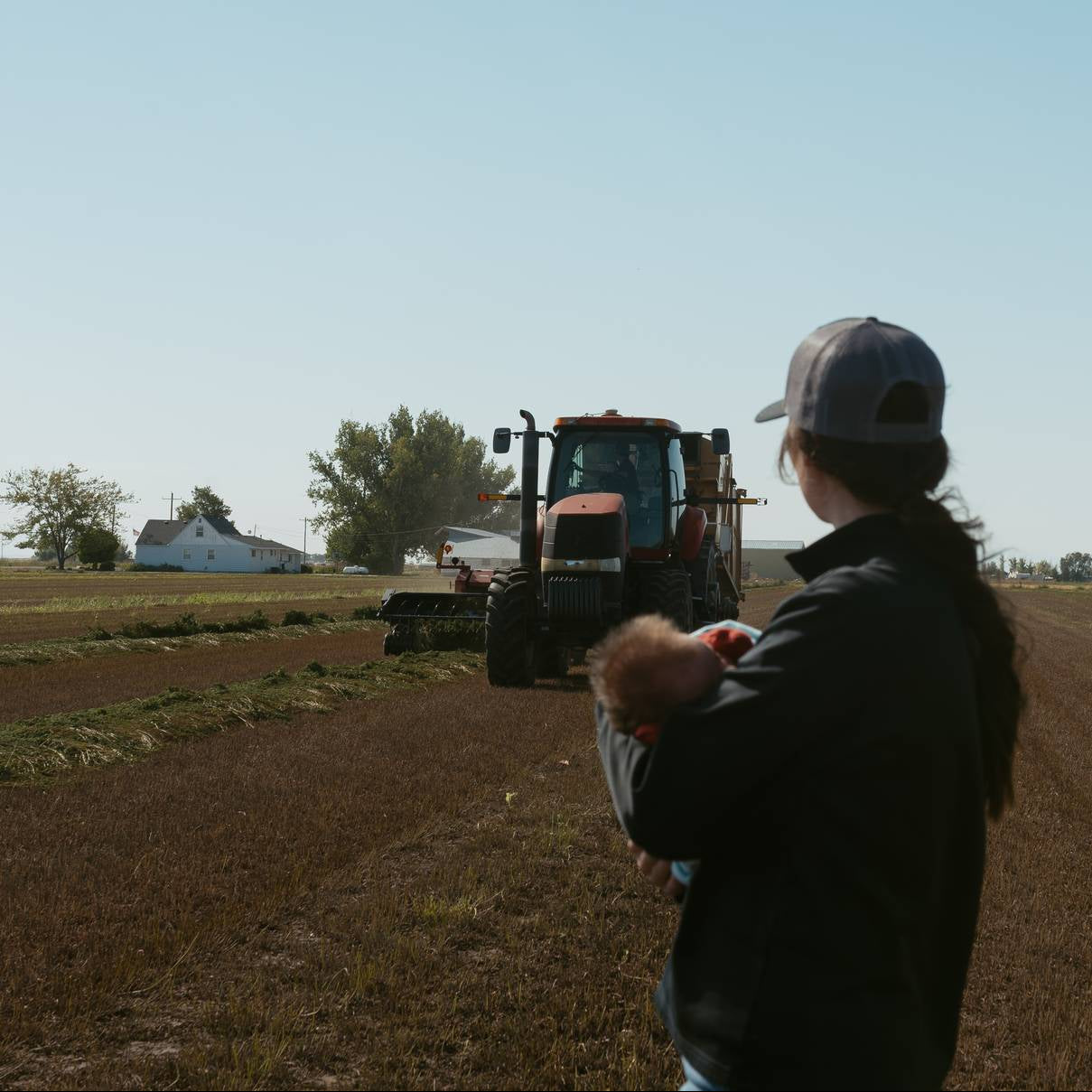 Person holding a child in a field with a tractor in the background